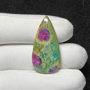 May include: A teardrop-shaped gemstone with a polished surface. The stone features a vibrant mix of colours, including deep pink, teal, and green hues. The gemstone's pattern includes circular pink inclusions, creating a unique design. The stone is held by a gloved hand.