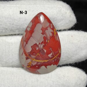 May include: A polished, teardrop-shaped gemstone. The stone displays a pattern of red, grey, and yellow veins. The text "N-3" is visible. The gemstone is held by a gloved hand.