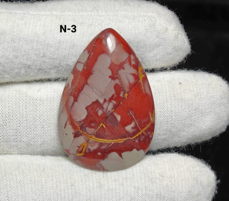 May include: A polished, teardrop-shaped gemstone. The stone displays a pattern of red, grey, and yellow veins. The text "N-3" is visible. The gemstone is held by a gloved hand.