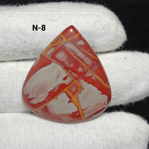 May include: A polished, teardrop-shaped gemstone with a striking pattern of red, white, and yellow veins. The text "N-8" is visible. The gemstone is displayed on white fabric. A unique piece for jewellery making.