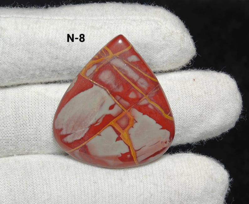 May include: A polished, teardrop-shaped gemstone with a striking pattern of red, white, and yellow veins. The text "N-8" is visible. The gemstone is displayed on white fabric. A unique piece for jewellery making.