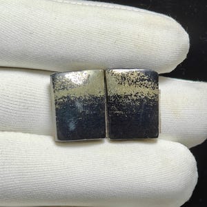 May include: A pair of rectangular cufflinks, each with a dark base and a speckled, gold-coloured top. The cufflinks are presented against a white fabric background, likely for display purposes. A jewellery or accessory item.