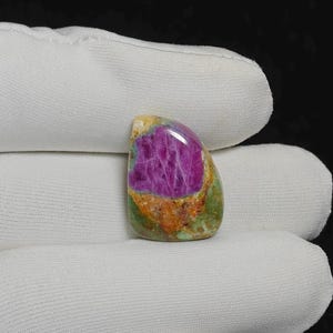 Pode incluir: Uma pedra preciosa polida com um vibrante centro magenta, rodeada por uma faixa de tons laranja e verde. A pedra tem uma superfície lisa e brilhante e uma forma irregular e arredondada. A pedra preciosa é segurada em um tecido branco.