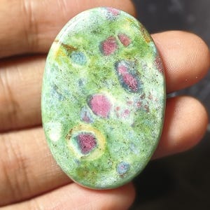 May include: An oval-shaped cabochon gemstone featuring a polished surface with a mottled light green base color. The stone is speckled with distinct, embedded deep pink and magenta inclusions, along with subtle hints of blue and yellow mineral deposits.