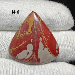 May include: A triangular, polished gemstone with a red base colour, featuring white and yellow veining. The stone has a smooth surface and is held by a gloved hand. The text "N-6" is visible in the upper left corner. This is a natural stone.