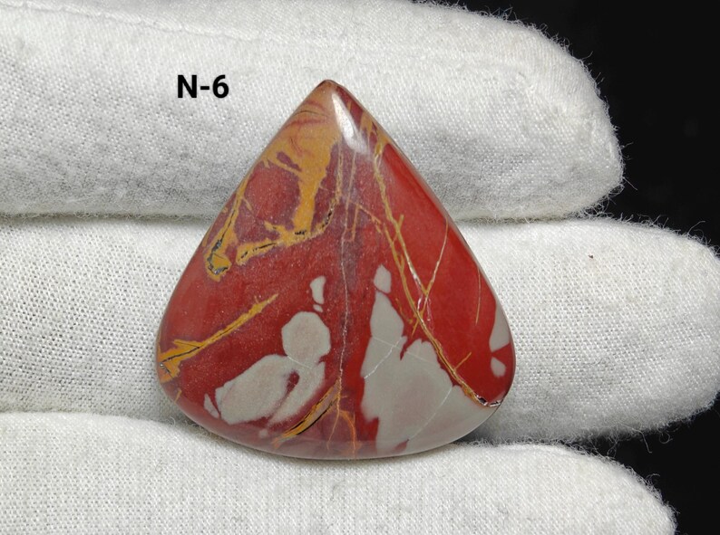 May include: A triangular, polished gemstone with a red base colour, featuring white and yellow veining. The stone has a smooth surface and is held by a gloved hand. The text "N-6" is visible in the upper left corner. This is a natural stone.