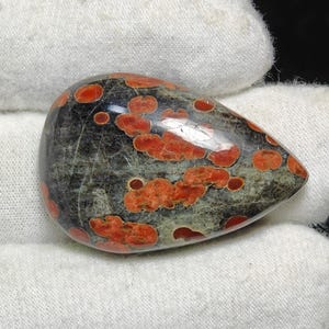 May include: A teardrop-shaped cabochon of red and black jasper with a speckled pattern. The stone is polished and has a smooth surface.