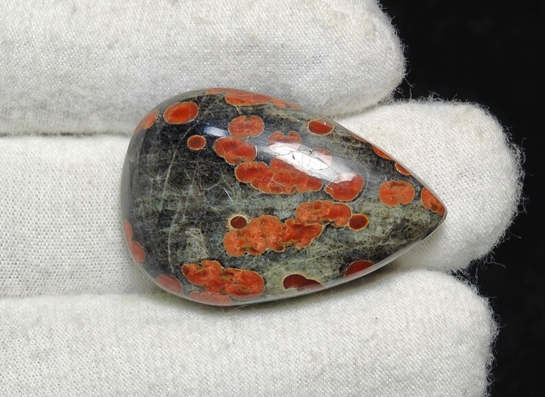 May include: A teardrop-shaped cabochon of red and black jasper with a speckled pattern. The stone is polished and has a smooth surface.