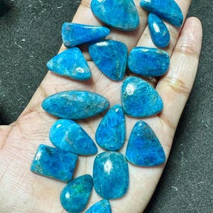 May include: A selection of polished, bright blue gemstones in diverse shapes, such as oval, teardrop, and rectangular. The stones are displayed in a hand, highlighting their natural textures and colour variations.
