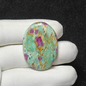 Puede incluir: Gema ovalada con una vibrante mezcla de colores. La piedra presenta una base verde claro con vetas marrones y naranjas. Incrustados hay parches de rosa intenso y rojo, creando un contraste llamativo. La superficie pulida refleja la luz.