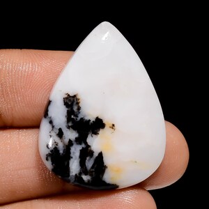 May include: White cabochon gemstone with black and brown dendritic inclusions. The gemstone is shaped like a teardrop and has a smooth, polished surface.