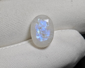 Könnte beinhalten: Ovale, schillernder Mondstein-Edelstein. Der Stein hat eine milchig-weiße Grundfarbe mit blauen Reflexen und eine facettierte Oberfläche. Der Edelstein wird auf weißem Stoff präsentiert.