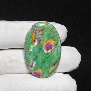Peut inclure: Pierre précieuse ovale avec une base verte vibrante, parsemée d'inclusions magenta et jaunes. La pierre polie est présentée sur un fond de tissu blanc, mettant en valeur ses motifs et textures naturels. La combinaison de couleurs unique de la pierre précieuse est frappante.