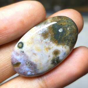 May include: An oval-shaped gemstone with a polished surface. The stone features a mix of colors, including white, brown, green, and purple. The gemstone is held in a hand, showcasing its natural beauty and unique patterns. A small, circular pattern is visible on the stone.