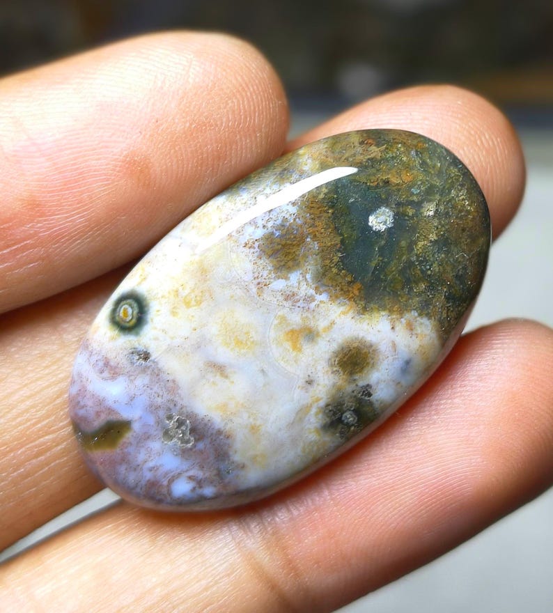 May include: An oval-shaped gemstone with a polished surface. The stone features a mix of colors, including white, brown, green, and purple. The gemstone is held in a hand, showcasing its natural beauty and unique patterns. A small, circular pattern is visible on the stone.