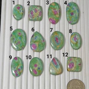 May include: A collection of twelve polished gemstones with a mix of shapes, including ovals, teardrops, and rectangles. The stones feature a green base color with pink and yellow inclusions. A US quarter is included for size reference.