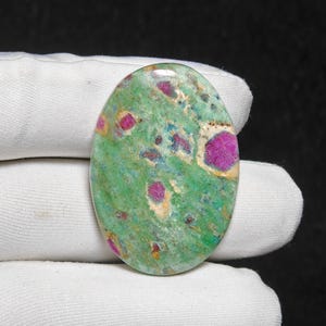 Puede incluir: Gema ovalada con una base verde salpicada de vibrantes inclusiones rosas y rojas. La piedra tiene una superficie pulida, y las inclusiones varían en tamaño y forma, creando un patrón único. La gema se presenta sobre un fondo blanco.