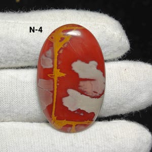 May include: Polished oval gemstone with a red base, white and yellow patterns, and the text "N-4". The stone is held by a gloved hand. The gemstone's surface is smooth and reflective, showcasing natural variations in colour and pattern.