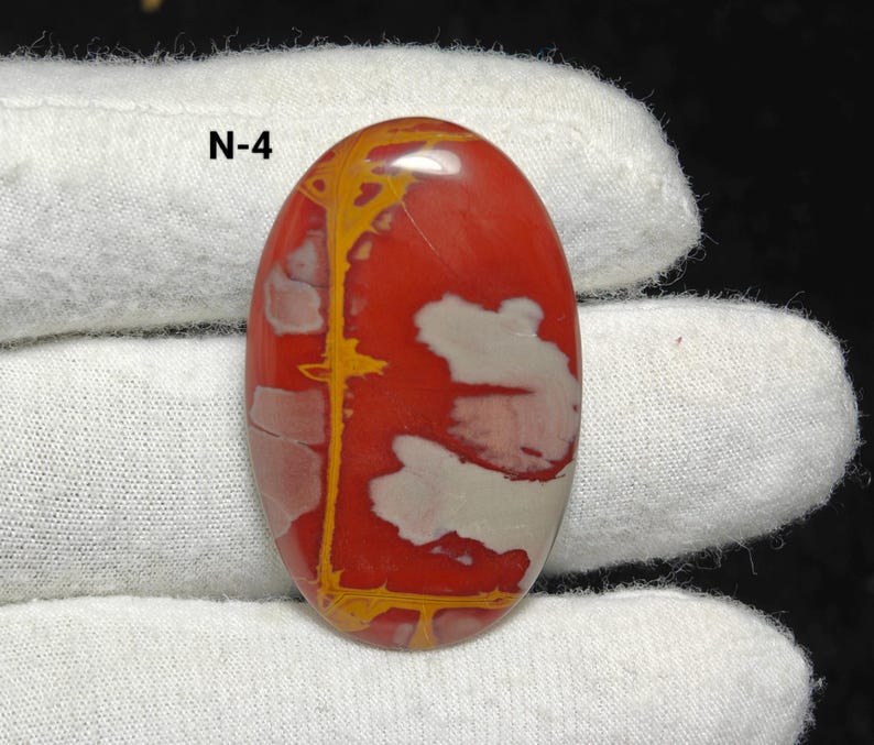 May include: Polished oval gemstone with a red base, white and yellow patterns, and the text "N-4". The stone is held by a gloved hand. The gemstone's surface is smooth and reflective, showcasing natural variations in colour and pattern.