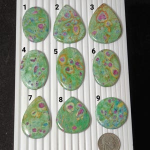 Puede incluir: Nueve piedras preciosas pulidas de rubí zoisita en varias formas, incluyendo ovaladas, de lágrima y redondas. Las piedras presentan un color base verde con inclusiones rosas y naranjas. Se incluye una moneda de veinticinco centavos como referencia de tamaño.