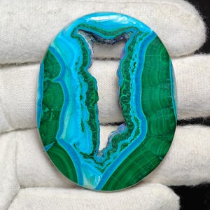 Puede incluir: Una piedra preciosa ovalada pulida con un gran agujero en el centro. La piedra preciosa tiene un patrón en capas de colores verde y azul.