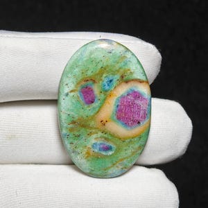 Könnte beinhalten: Ein ovaler Edelstein mit grüner Grundfarbe, gesprenkelt mit dunkleren Grün- und Brauntönen. Er hat leuchtend magenta- und blaue Einschlüsse und einen cremefarbenen Rand. Der Stein wird von einem weißen Handschuh gehalten.