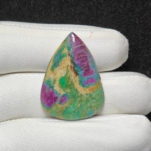 Puede incluir: Una gema en forma de lágrima con una mezcla de colores. La piedra presenta áreas de magenta intenso, verde azulado y verde, con secciones más claras de color beige y crema. La piedra es sostenida por una mano enguantada de blanco.