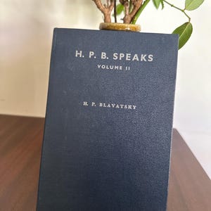 Könnte beinhalten: Ein marineblaues Buch mit dem Titel "H. P. B. Speaks Volume II" von H. P. Blavatsky. Das Buch steht auf einer Holzoberfläche, mit einer kleinen Pflanze in einem Keramiktopf dahinter. Der Text ist in silbernen Buchstaben.