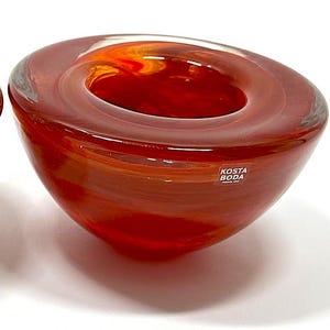 Op de afbeelding: Twee rode glazen kommen met een cirkelvormige opening. De kommen hebben een gelaagde, gemarmerde uitstraling met tinten oranje en rood. Elke kom heeft een label met de tekst "KOSTA BODA" en "napolitano gioielli BARLETTA".