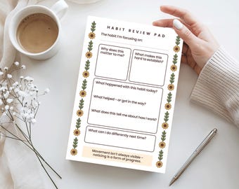 May include: A habit review pad with a floral border, featuring prompts for self-reflection. The pad includes questions like "Why does this matter to me?" and "What can I do differently next time?" A cup of coffee, flowers, and a pen are also in the image.