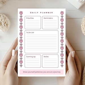 May include: A white daily planner with pink floral detailing and the words "DAILY PLANNER." The planner has sections for priorities, reminders, a to-do list, and notes. The bottom of the planner reads "Show yourself patience; you are on a journey."