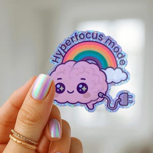 May include: A holographic sticker featuring a cartoon brain with a rainbow and the words "hyperfocus mode." The brain is light purple with a smiling face and a plug. The sticker has a glittery border.