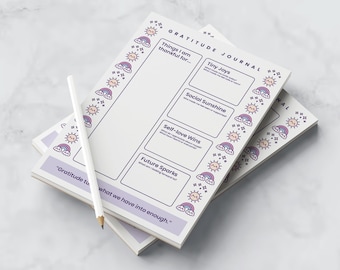 Puede incluir: Una pila de diarios de gratitud con un lápiz blanco. Los diarios tienen un fondo blanco con un borde lavanda y texto. El borde presenta soles, arcoíris y estrellas. El texto incluye "Diario de gratitud" y preguntas.