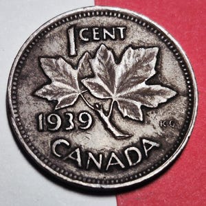 May include: A silver Canadian one-cent coin from 1939. The coin features two maple leaves, the text "1 CENT", the year "1939", and the word "CANADA". The coin has a detailed, textured surface.