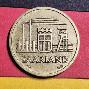 May include: A bronze-colored coin featuring a detailed relief of industrial structures, including a tower and a building with a shield. The word "SAARLAND" is inscribed at the bottom. The coin is set against a backdrop of red, black, and yellow.