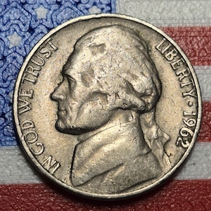 May include: A close-up of a 1962 nickel coin. The coin features a profile of Thomas Jefferson, with the words "IN GOD WE TRUST" and "LIBERTY" visible. The year "1962" is also displayed. The coin is silver in color.
