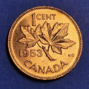 May include: A 1953 Canadian one-cent coin. The coin is gold-colored and features two maple leaves, the text "1 CENT", the year "1953", and the word "CANADA". The coin has a textured edge.