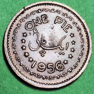 May include: A silver-colored coin with the text "ONE PIE" and the year "1956". The coin has an ornate border and several star-shaped designs. The coin's surface shows signs of wear.