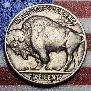 May include: A silver United States Buffalo nickel coin. The coin features a detailed image of a bison on one side and the words "FIVE CENTS" at the bottom. The words "UNITED STATES OF AMERICA" are at the top.