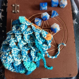 May include: A blue and white crocheted dice bag with a drawstring closure, sitting on a brown leather journal with a pentagram design. Six blue and white dice with gold numbers are scattered around the bag.