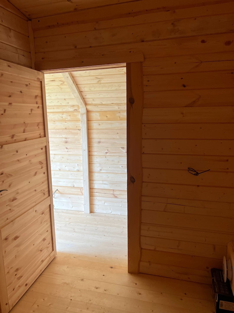 Puede incluir: Una puerta de madera abierta revela una habitaci&oacute;n con suelo y paredes de madera. La habitaci&oacute;n est&aacute; vac&iacute;a, excepto por algunos muebles.