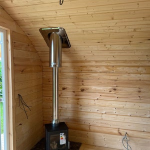 Puede incluir: Una estufa de le&ntilde;a negra con un tubo de chimenea de acero inoxidable y una base de azulejos negros. La estufa est&aacute; instalada en una esquina de una habitaci&oacute;n con paneles de madera en las paredes.