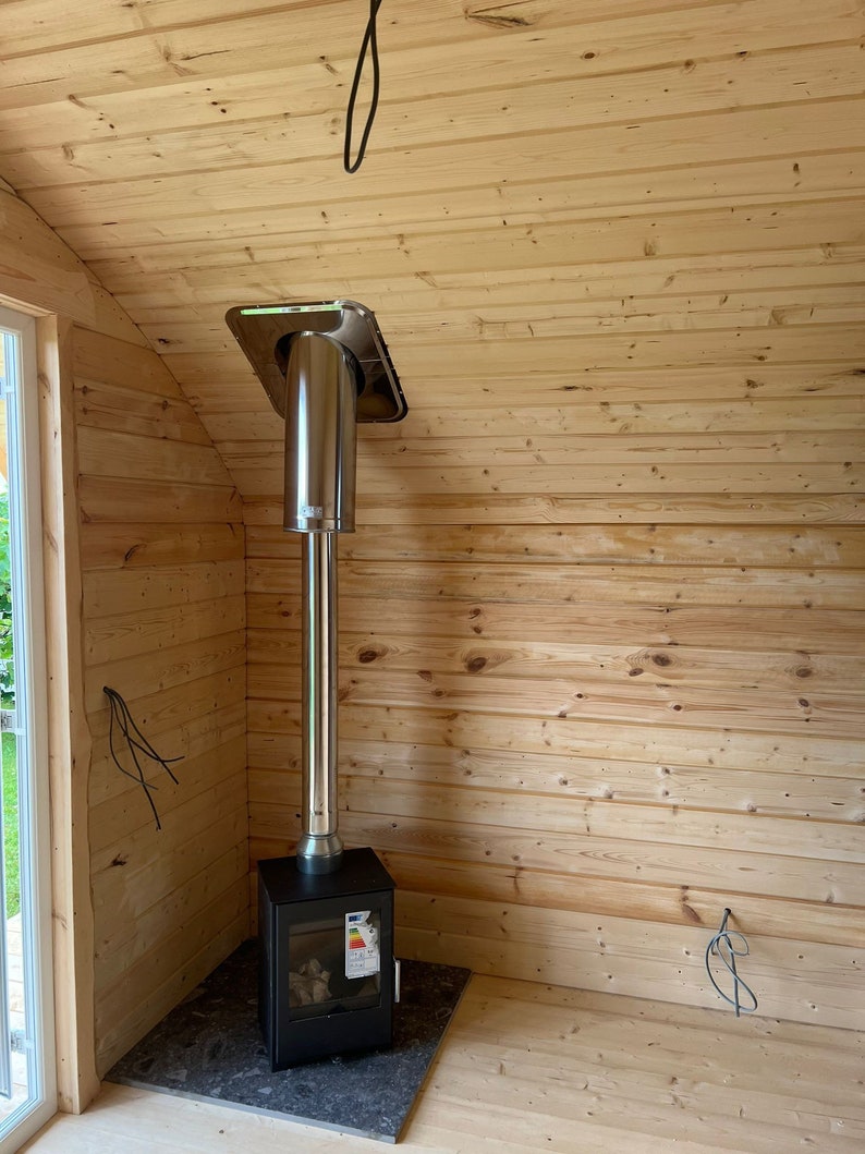 Puede incluir: Una estufa de le&ntilde;a negra con un tubo de chimenea de acero inoxidable y una base de azulejos negros. La estufa est&aacute; instalada en una esquina de una habitaci&oacute;n con paneles de madera en las paredes.