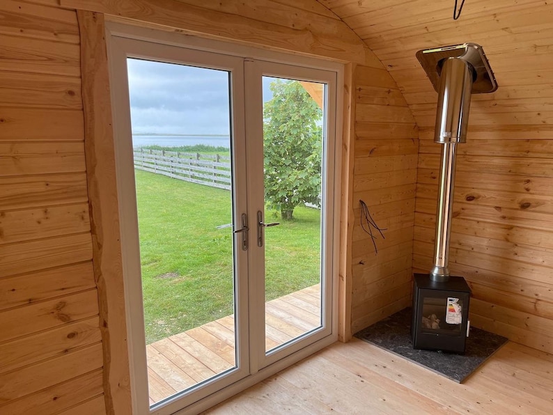 Puede incluir: Una peque&ntilde;a caba&ntilde;a de madera con una estufa de le&ntilde;a y vistas a un campo de hierba y agua. La caba&ntilde;a tiene una ventana grande con marco blanco y una puerta de madera.