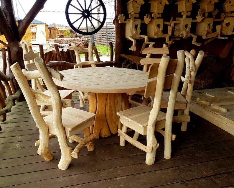 May include: A light-colored wooden outdoor dining set. The set includes a round table and several chairs, all crafted from natural-looking wood. The chairs have unique branch-like supports. The table and chairs are on a wooden deck.