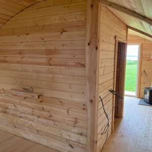 Puede incluir: Una peque&ntilde;a caba&ntilde;a de madera con una estufa de le&ntilde;a y una ventana con vistas a un campo de hierba. La caba&ntilde;a est&aacute; hecha de madera natural y tiene un techo curvo.
