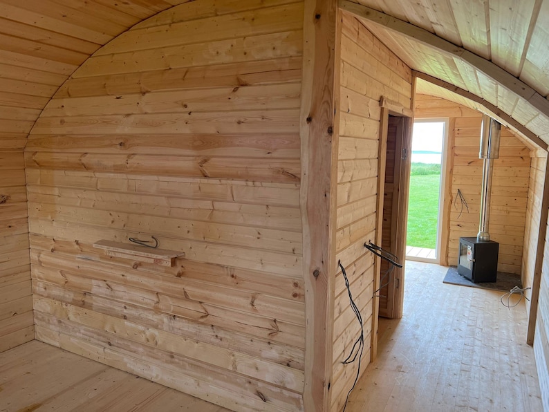 Puede incluir: Una peque&ntilde;a caba&ntilde;a de madera con una estufa de le&ntilde;a y una ventana con vistas a un campo de hierba. La caba&ntilde;a est&aacute; hecha de madera natural y tiene un techo curvo.