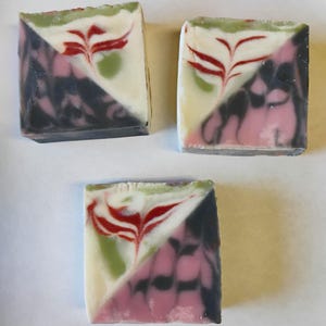 May include: Three square handmade soaps with a diagonal color split. One side is white with a red floral design, green and red accents. The other side is black and pink. The soaps are on a white surface.
