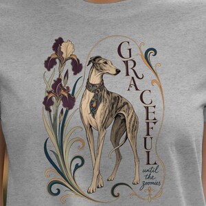 May include: A heather gray t-shirt featuring a graphic illustration of a brindle greyhound standing next to dark purple iris flowers. The design includes the text "GRACEFUL" in a vertical orientation and the smaller script text "until the zoomies".
