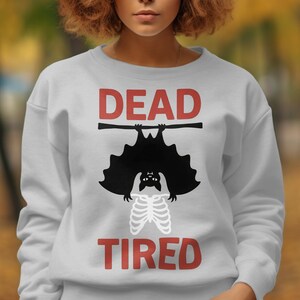 May include: A light gray crewneck sweatshirt featuring a graphic print of a black bat hanging from a horizontal bar. Below the bat is a white skeletal rib cage illustration. The red text "DEAD" appears above the bat and "TIRED" below it.
